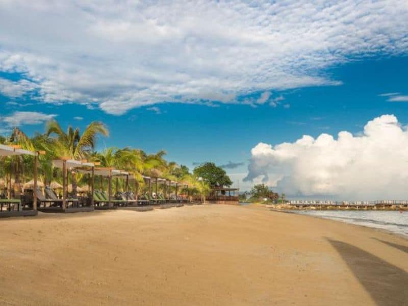 View of Iberostar Rose Hall, displaying its sandy beach, lush trees, clear waters, and quaint cottages.