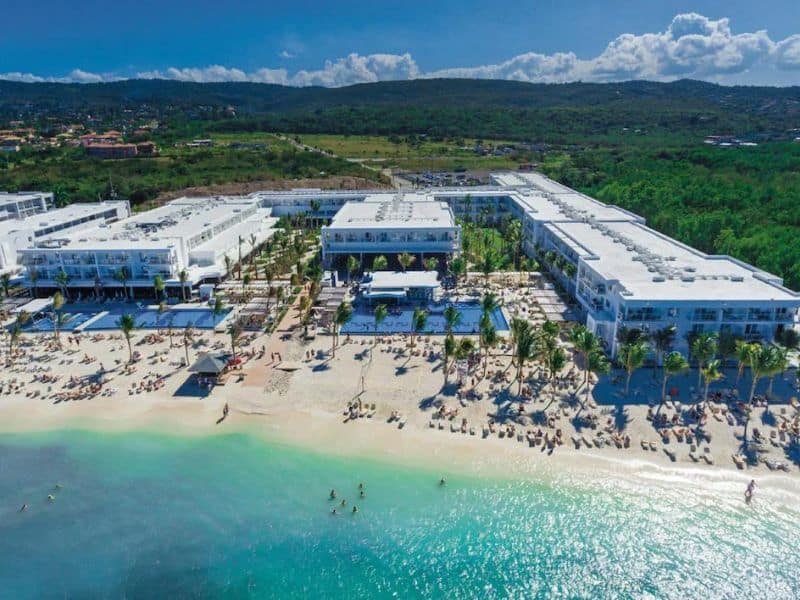 An aerial view of RIU Reggae Resort, featuring tall buildings, sandy beach, pools, palm trees, and people swimming along the shore.