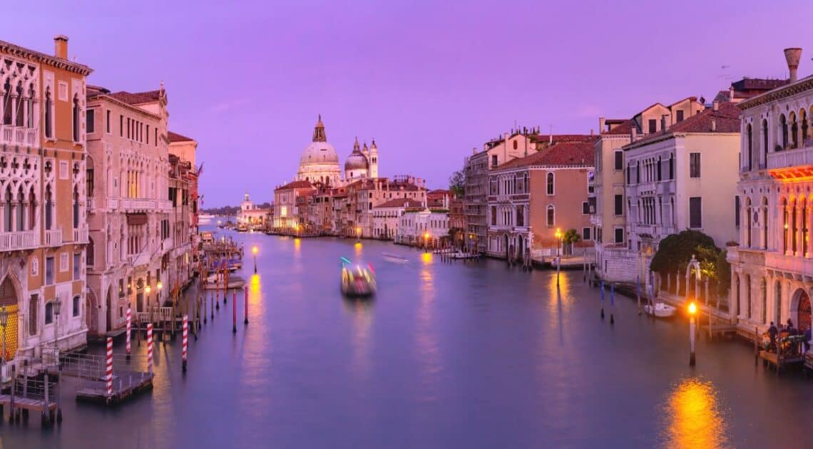 One of the 12 best Venice shore excursions. A stunning scene, with light purple sunlight casting a magical glow over the charming canals and gondolas. The city's beauty shines as the sun dips below the horizon, creating a serene and romantic ambiance.