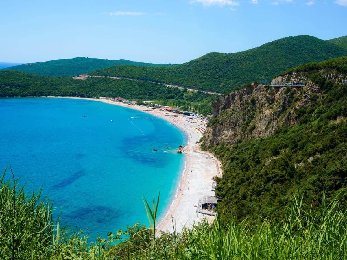 Jaz Beach on the Adriatic coast, a stretch of golden sands set against turquoise waters, backed by gentle hills, and frequented by sunbathers and swimmers seeking the Mediterranean sun.