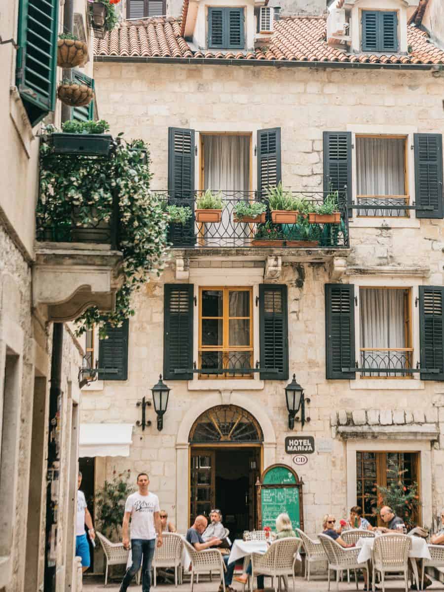 A cozy caf&eacute; in Kotor's old town, with patrons sipping coffee under the shadow of historic stone buildings, creating an ambiance of relaxation amidst the city's rich history.