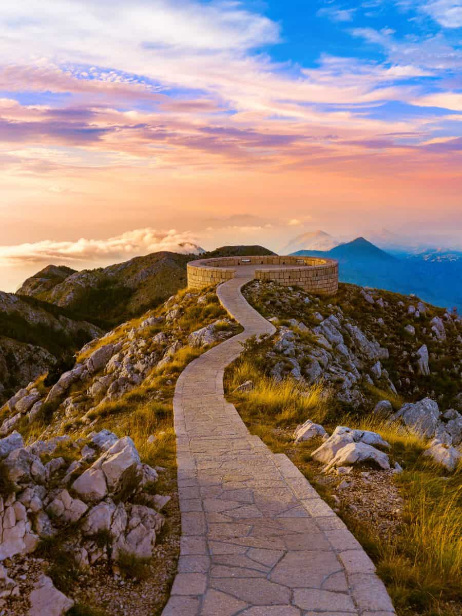 Lovćen National Park during sunset, where the sky turns orange and gold. The setting sun makes the tall mountains and trees look even more beautiful, giving everything a warm glow.