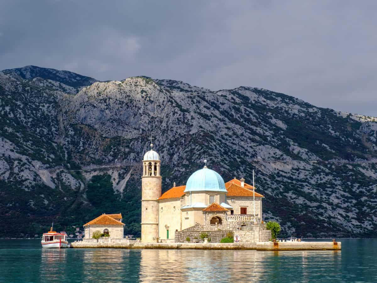Our Lady of the Rocks island in the Bay of Kotor, Montenegro, with its iconic white stone church showcasing a blue-domed roof, set against the backdrop of azure waters.