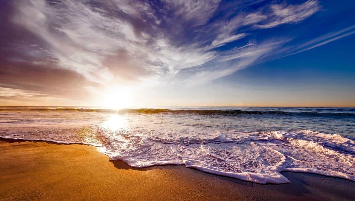 A serene beach scene at sunset with the sun casting a warm glow over the frothy waves as they gently wash onto the sandy shore, under a vast sky with wispy clouds.