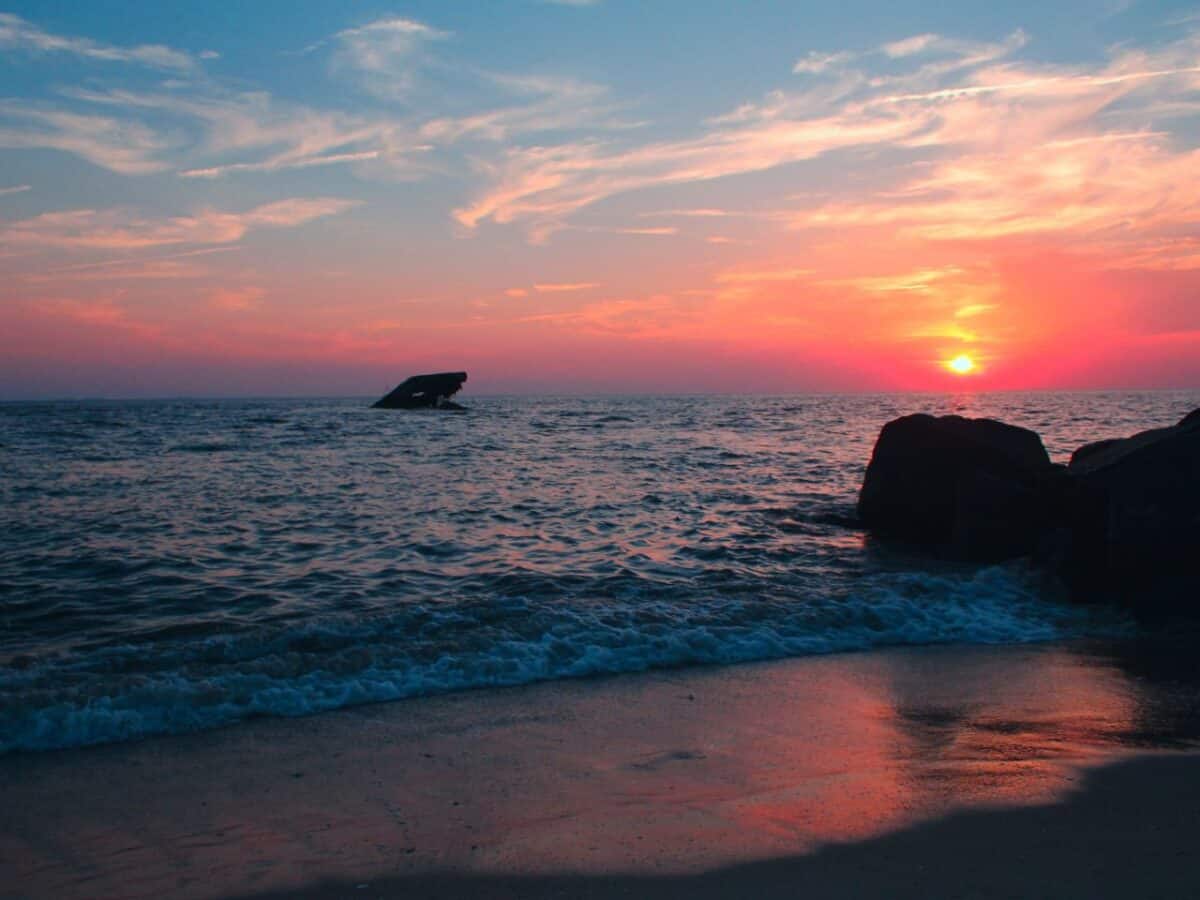 Twilight at Cape May with the sun setting on the horizon, illuminating the sky in shades of pink and orange, silhouetting a shipwreck on the right against the tranquil sea.