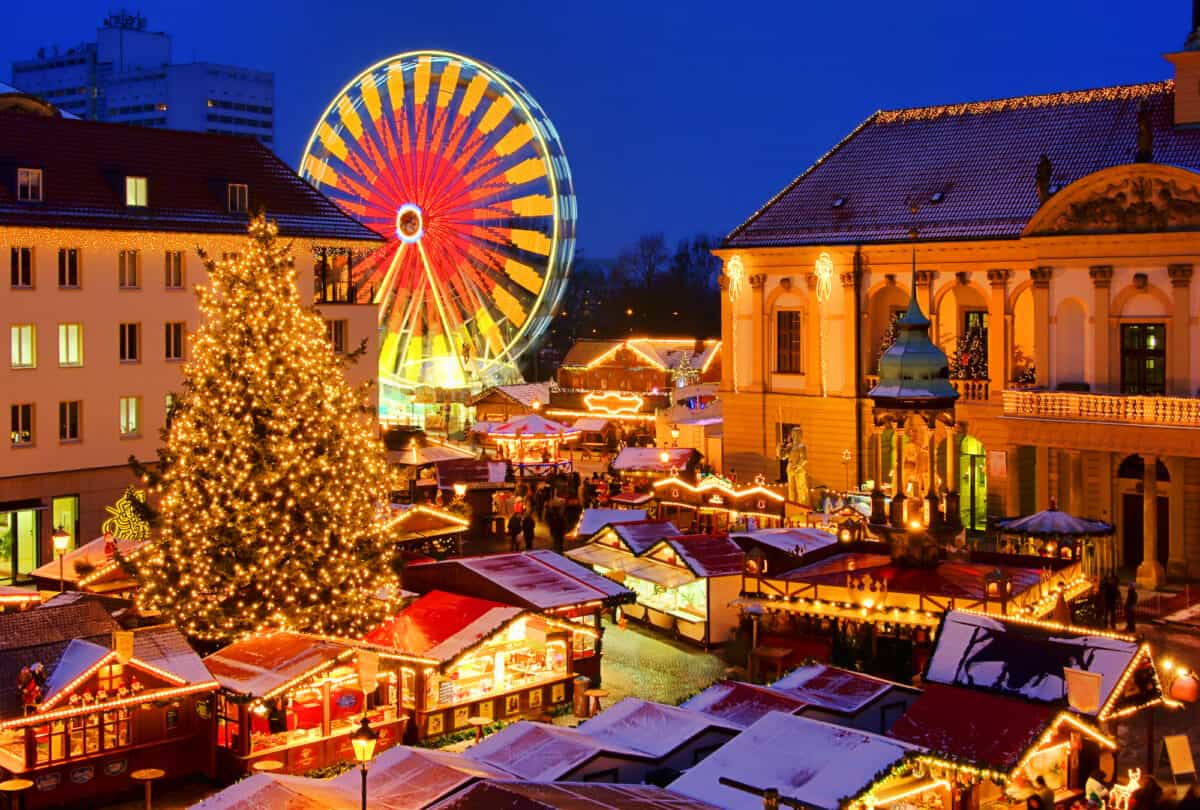 A vibrant Christmas market with a large Ferris wheel illuminated in blue and yellow lights, surrounded by festively decorated stalls and a snow-covered Christmas tree.
