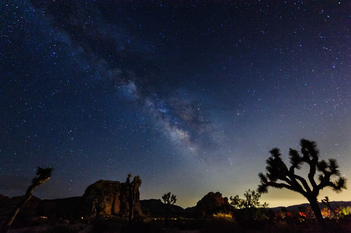 A panoramic night sky view over Joshua Tree National Park, showcasing a vibrant Milky Way stretching across the horizon, silhouetting distinctive Joshua trees and rock formations