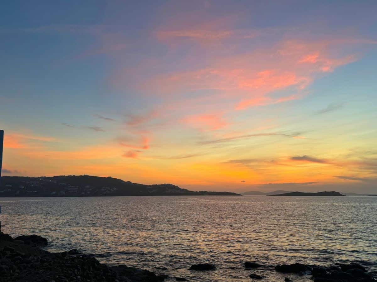 An ocean view of Mykonos, Greece during sunset, with vibrant hues in the sky and the serene Aegean Sea in the foreground.