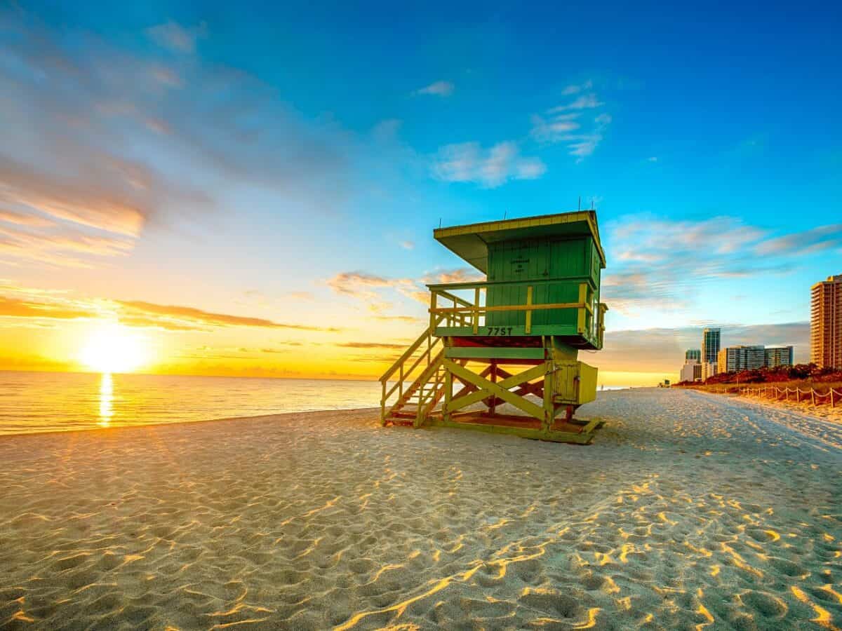 A lifeguard tower in South Beach, Miami, painted in green and sitting on the sand against a backdrop of a stunning sunrise, with the ocean on one side and the city skyline on the other.