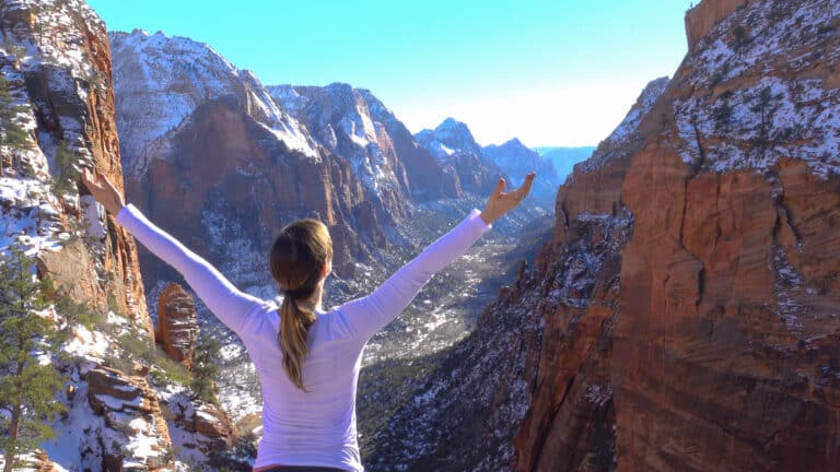 Woman hiking on a trail in Zion National Park with sparse snow on the ground and towering red rock formations in the background.