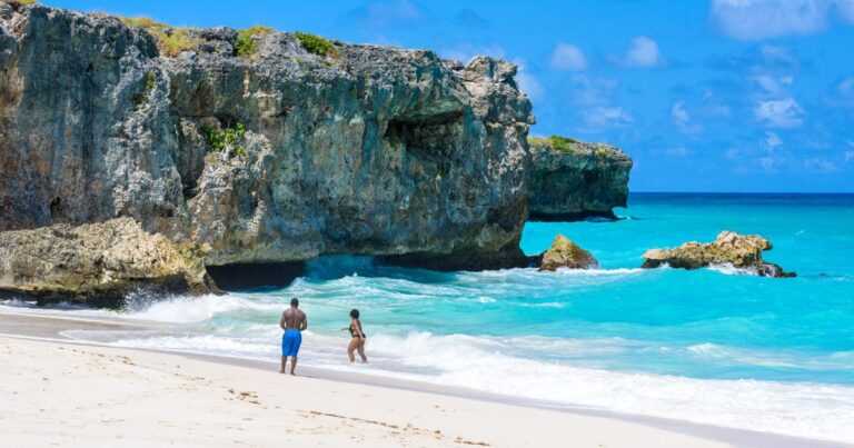 A serene beach in Barbados with clear blue waters, white sands, and giant rocks in the background with couples playing in the waves.