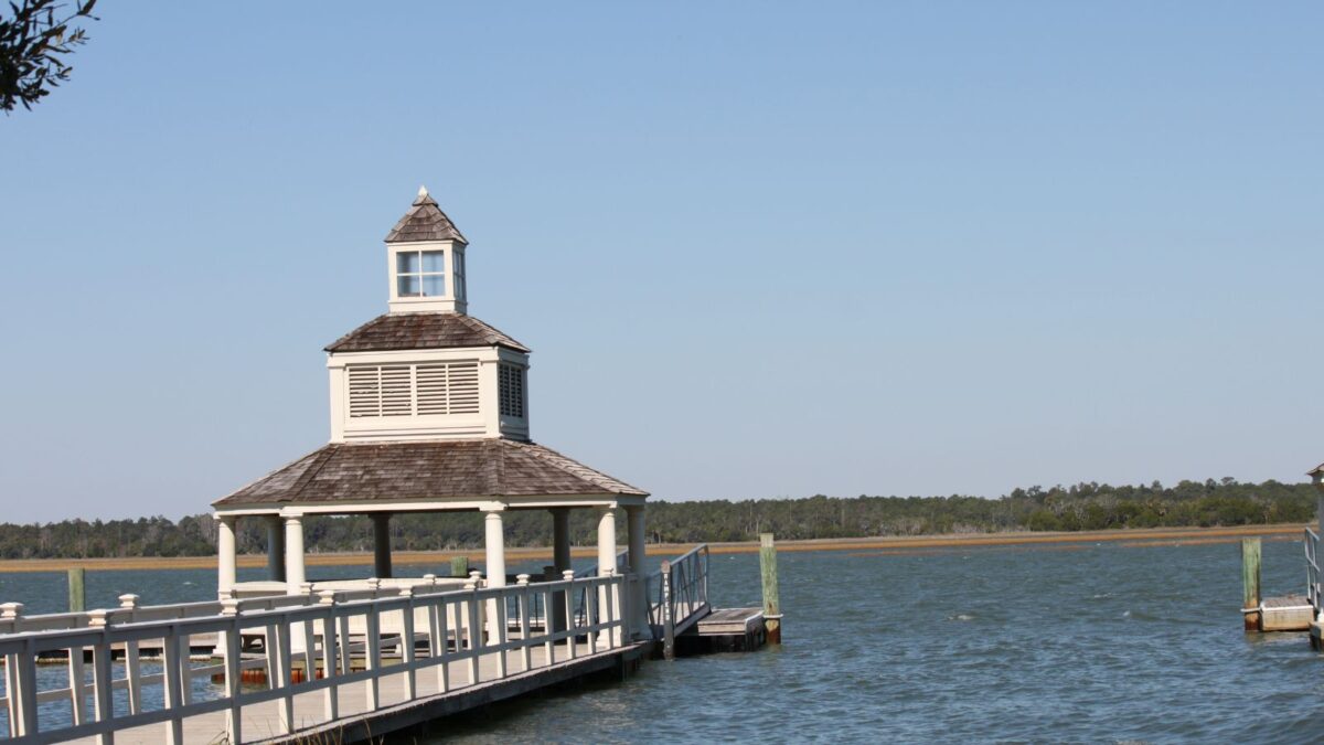 A gazebo with a pointed roof sits at the end of a wooden dock, overlooking a calm body of water. The sky is clear and blue, and the gazebo casts a crisp shadow on the white railing of the dock. The backdrop reveals a stretch of trees and clear skies.