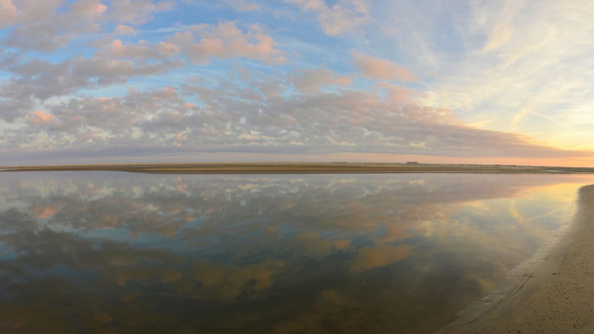 A panoramic view of a tranquil water body reflecting a sky with soft pink and blue hues of dawn or dusk, with a sandbar to the right.