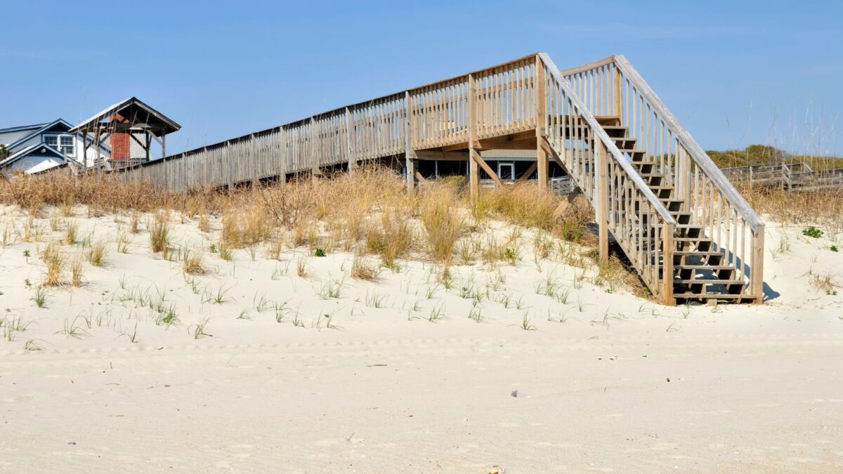 A wooden staircase over sand dunes leading to a beach house, with dry coastal grass and a clear blue sky in the background.