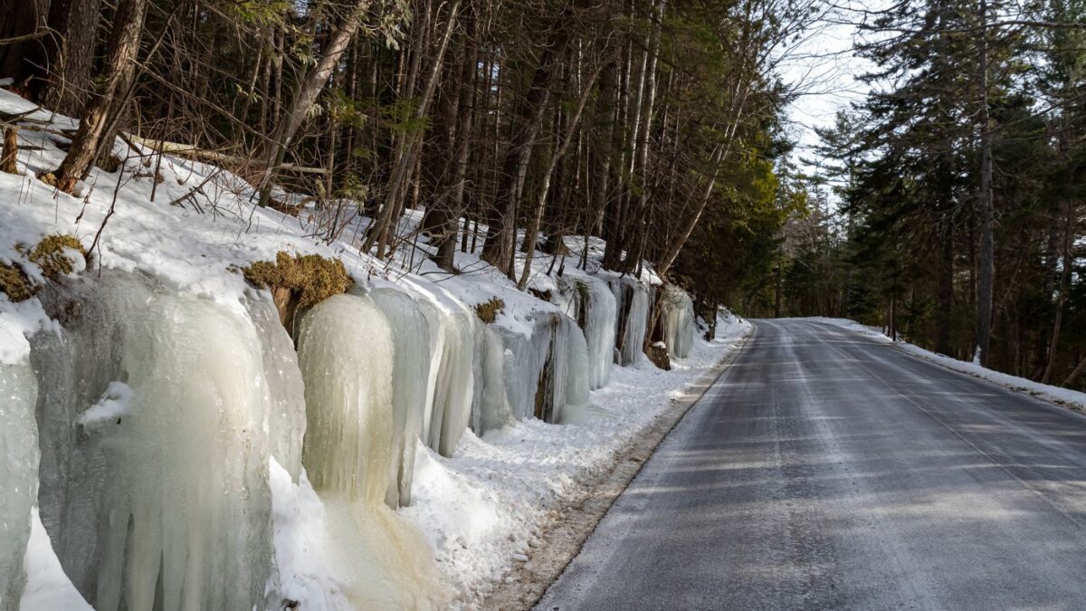 Icy roadside with icicles and snow-covered trees lining a clear road in a wintery forest setting.