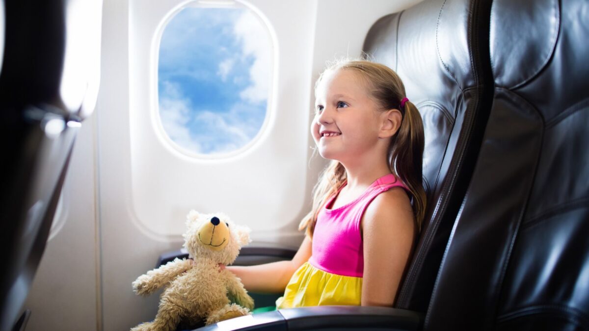 A young girl with blonde hair, wearing a pink and yellow dress, sits in an airplane seat, smiling and looking out the window. She is holding a teddy bear and appears to be enjoying her flight.