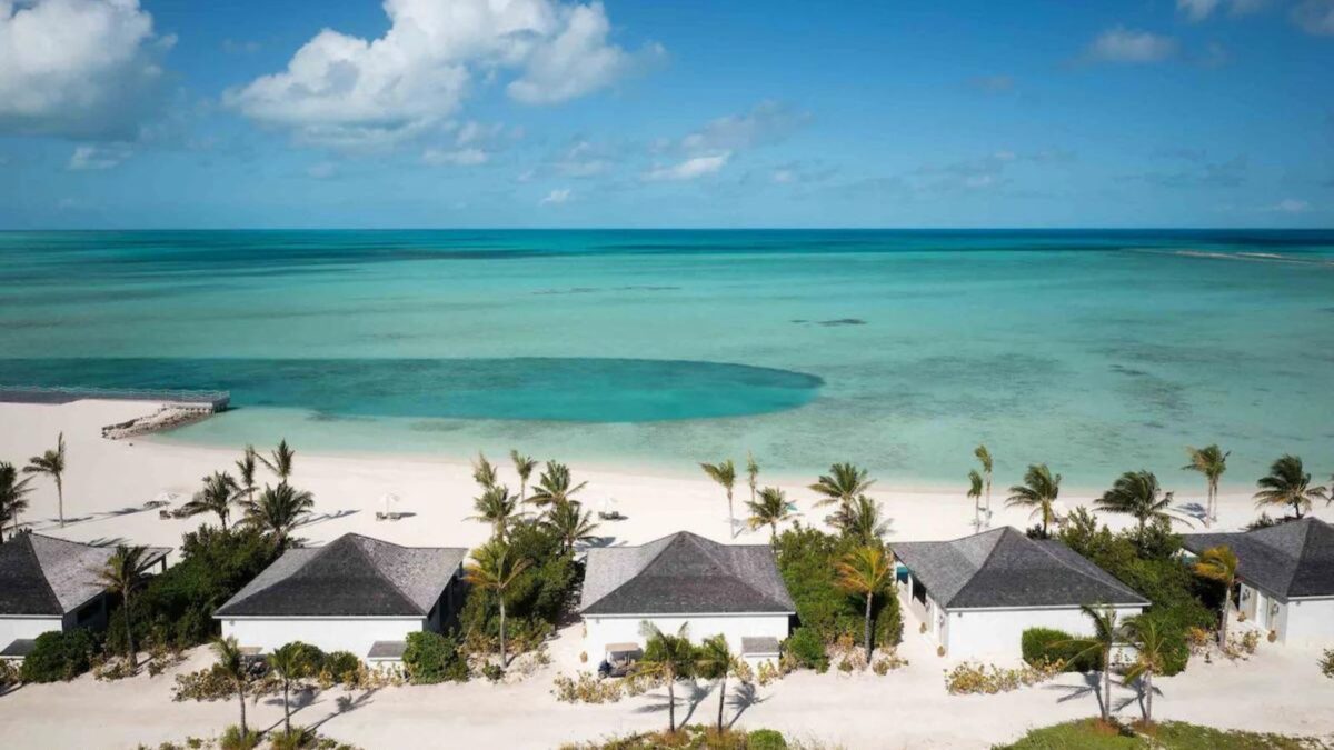 Beachfront villas with thatched roofs surrounded by palm trees on a white.sandy beach overlooking a turquoise sea.