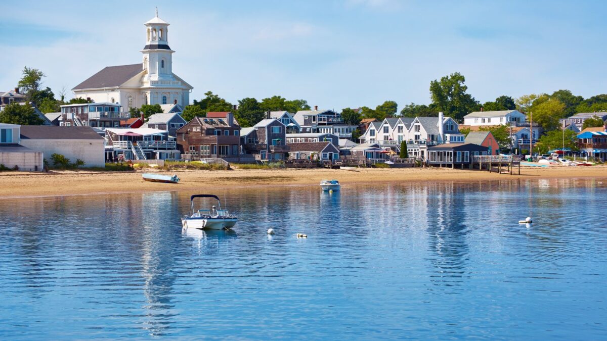 A picturesque view of Cape Cod, featuring a calm bay with boats and a backdrop of charming houses and a white church with a classic New England steeple.