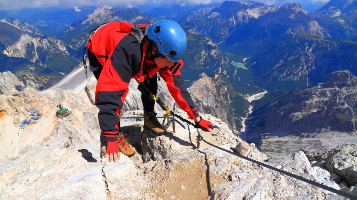 A climber in a red and black outfit with a blue helmet ascends a steep rocky path using a safety line, with a panoramic view of mountainous terrain in the background.