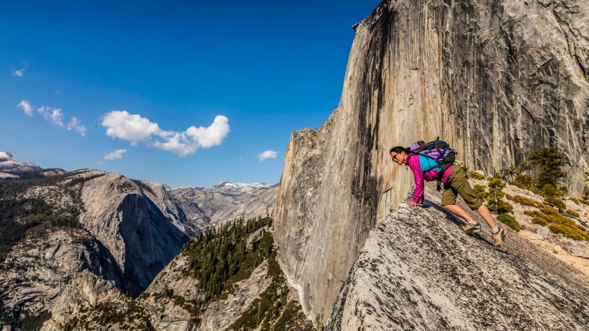 A hiker wearing a pink backpack leans into the slope of Half Dome, Yosemite, with a vast view of the valley behind her, illustrating the thrilling and exposed nature of this famous ascent.