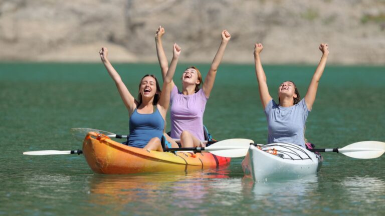 Three joyful women raise their arms in triumph while sitting in kayaks on a calm turquoise lake, celebrating an adventurous day out in nature.