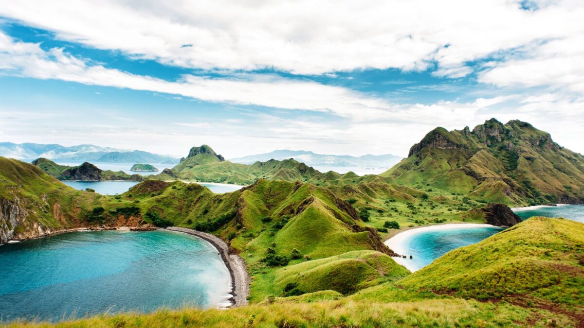 A panoramic view of the serene Komodo National Park, with its rolling green hills, calm blue bays, and distant rugged islands under a soft cloudy sky