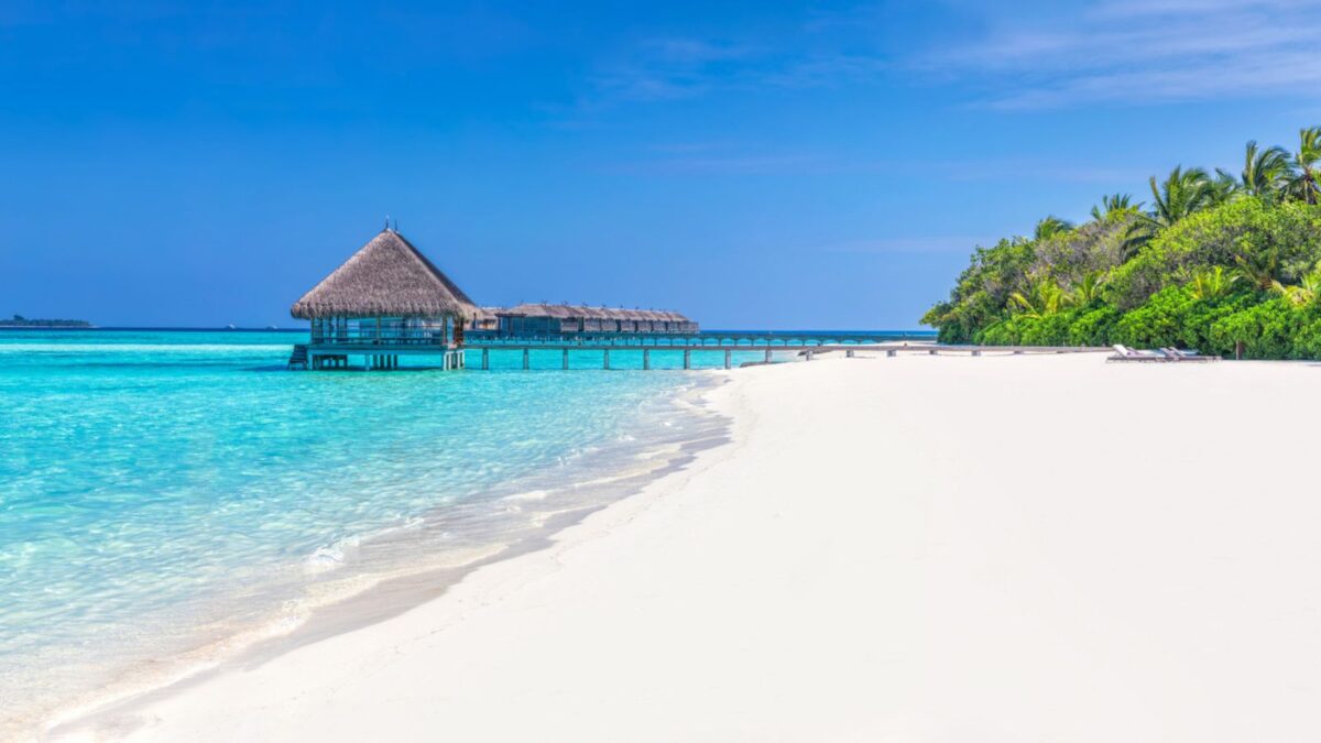 Expansive panoramic view of a Maldivian resort with overwater villas extending into the clear turquoise sea, white sandy beach, and lush tropical greenery