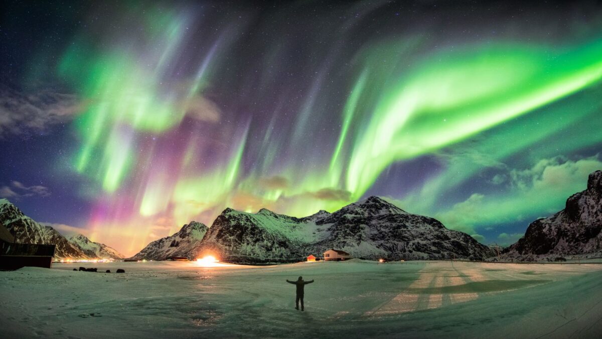 A person stands in awe beneath a vibrant display of Northern Lights over a snowy Norwegian landscape, with the aurora borealis casting a green and purple glow that illuminates the mountains and a small village in the distance.