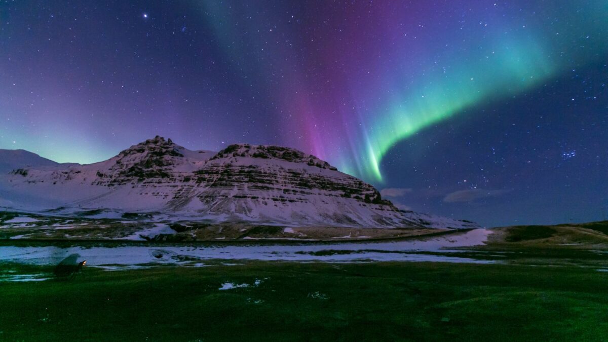 A magnificent aurora display arches over an Icelandic winter landscape, where the ground is sparsely covered with snow and the mountains in the background are under a gradient of green and purple night skies.