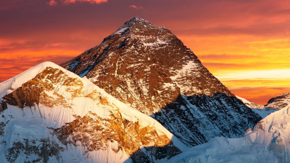 Majestic Mount Everest, bathed in the warm glow of sunset, with its peak touching the vibrant orange sky, while the surrounding snow-clad mountains bask in the last light of day