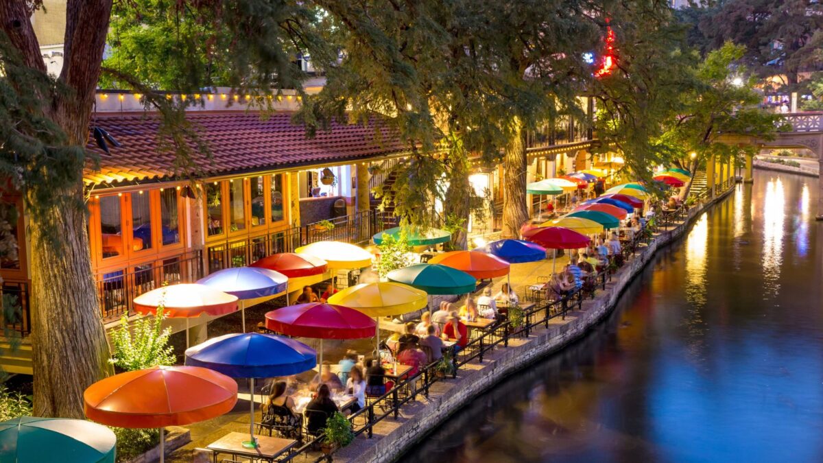 A vibrant evening scene along the San Antonio River Walk, with colorful umbrellas at outdoor cafes and diners enjoying the riverside ambiance.