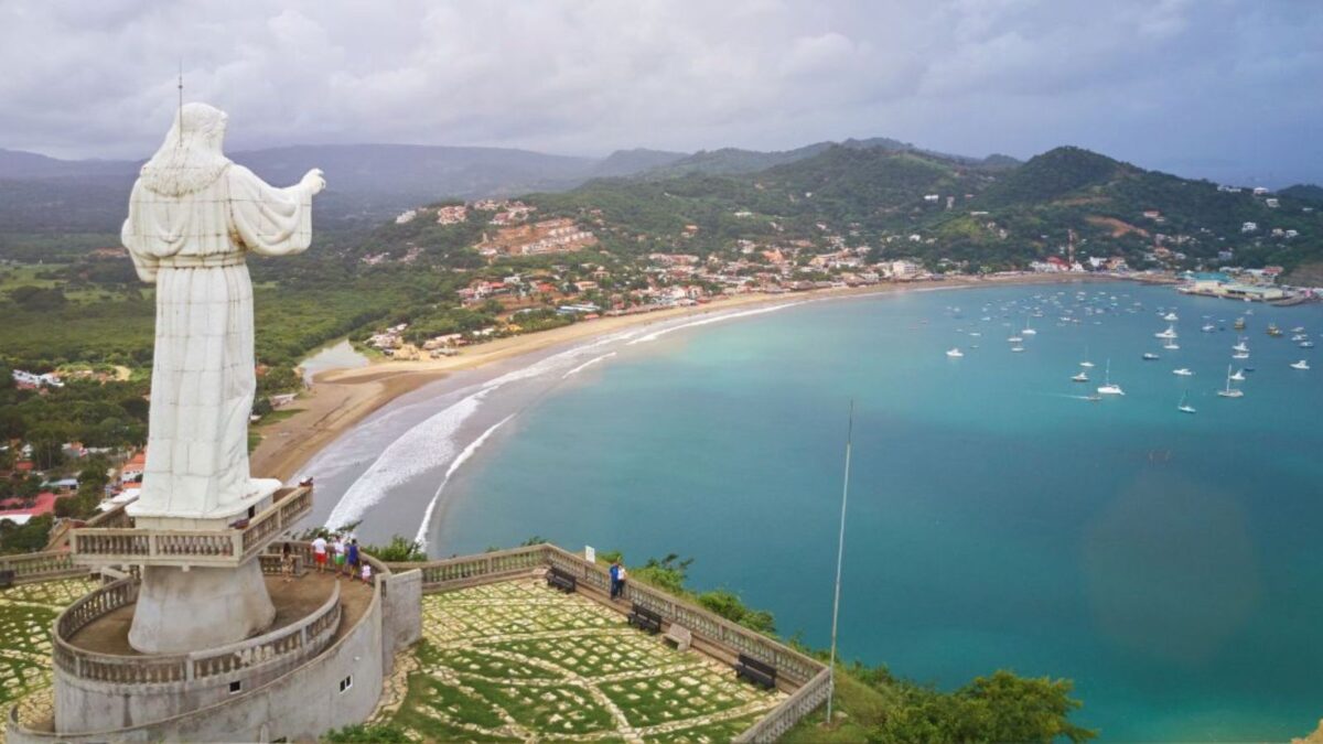 A towering white statue of Jesus overlooking San Juan del Sur bay, with a panoramic view of the beach and moored boats in the clear turquoise waters.