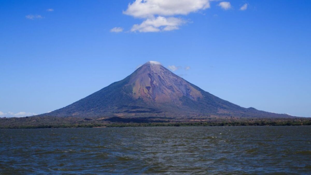 A majestic, conical volcano rising above a body of water, with a clear blue sky overhead.