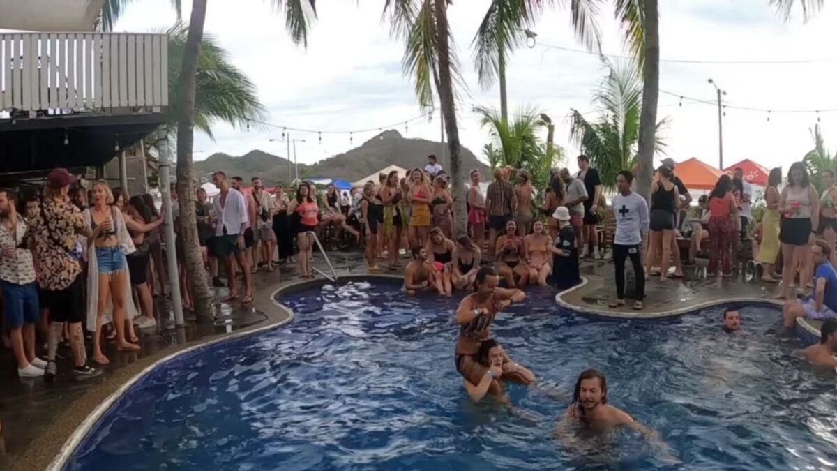 A lively pool party with a crowd gathered around a swim-up bar, set against a backdrop of palm trees and hills.