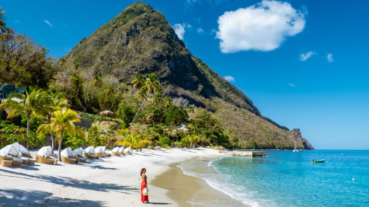 A picturesque tropical beach with a woman in a red dress walking along the shoreline, white sand cabanas in the foreground, and a lush green mountain in the background