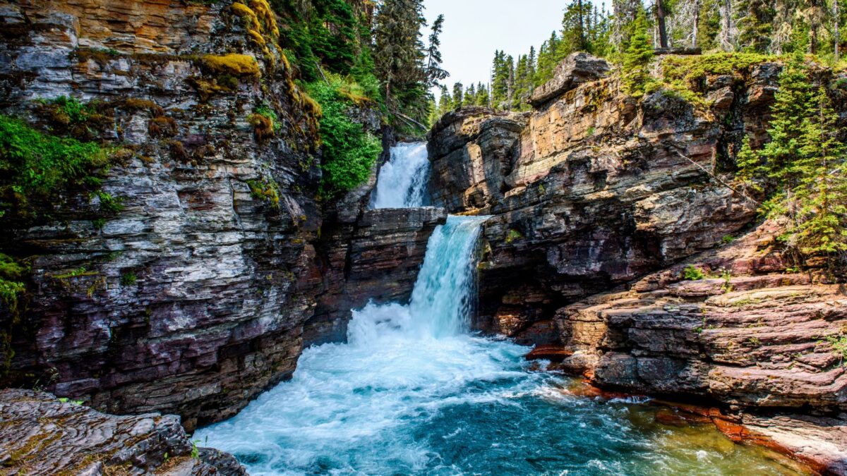A vibrant waterfall cascades down a rugged cliff with layered sedimentary rock, surrounded by lush greenery, into a turquoise pool below.