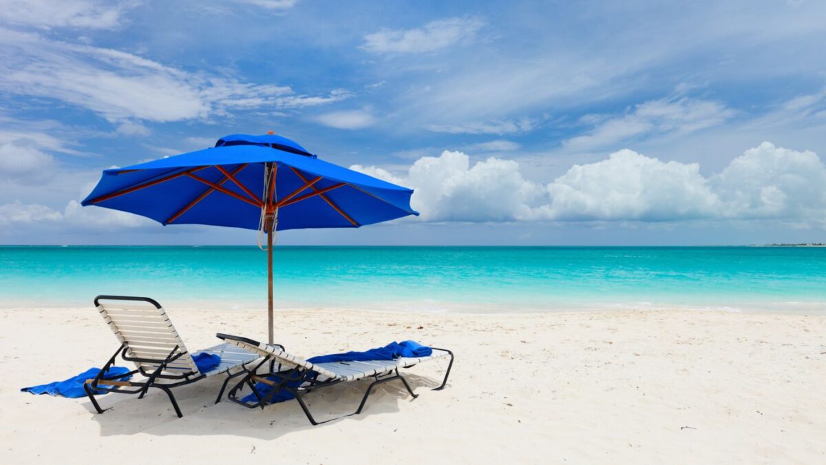 Inviting beach scene with a vibrant blue beach umbrella and two matching sun loungers on pristine white sand, overlooking a calm turquoise ocean under a sky dotted with fluffy clouds