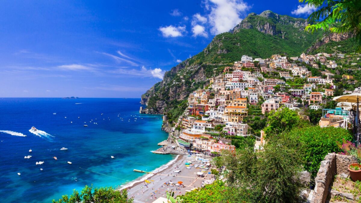 Panoramic view of the colorful buildings of Positano cascading down the hillside to a beach dotted with umbrellas, with boats dotting the azure sea."
