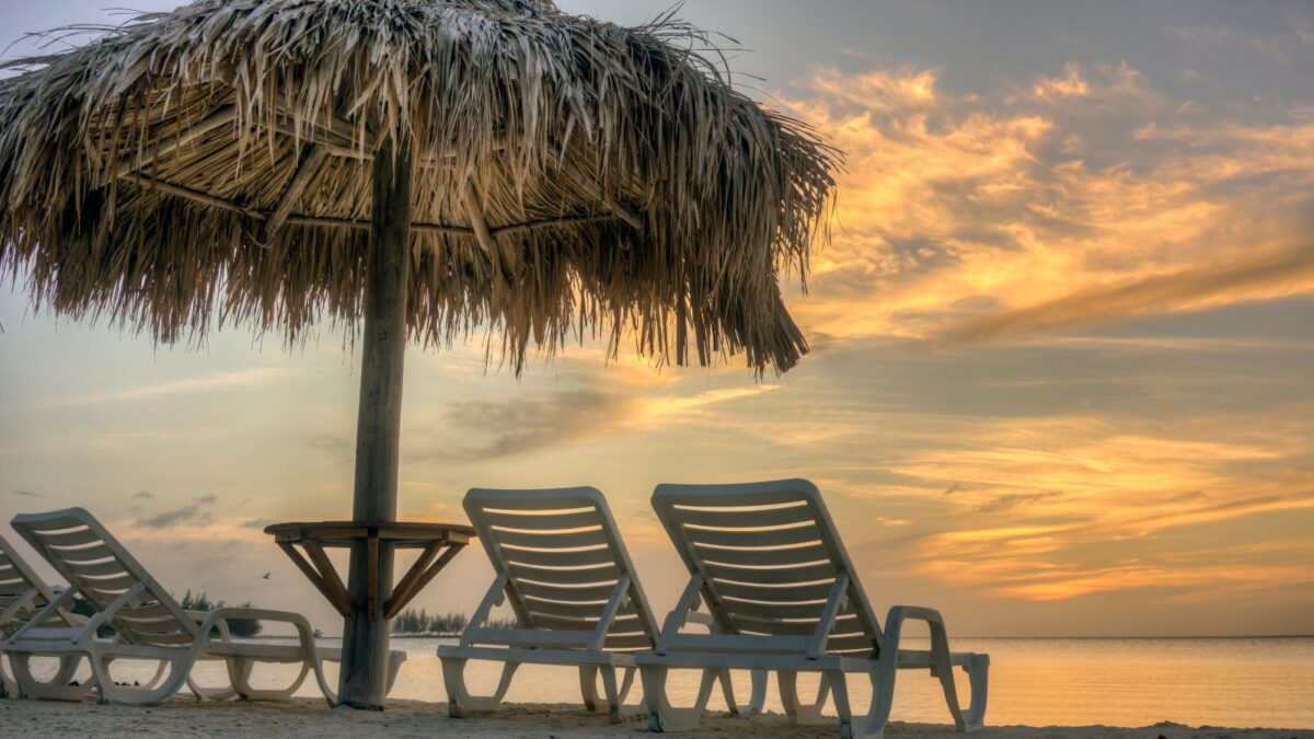 Best Roatan Honduras Beaches To Explore 11 Sunset at a beach with thatched umbrella and white lounge chairs.