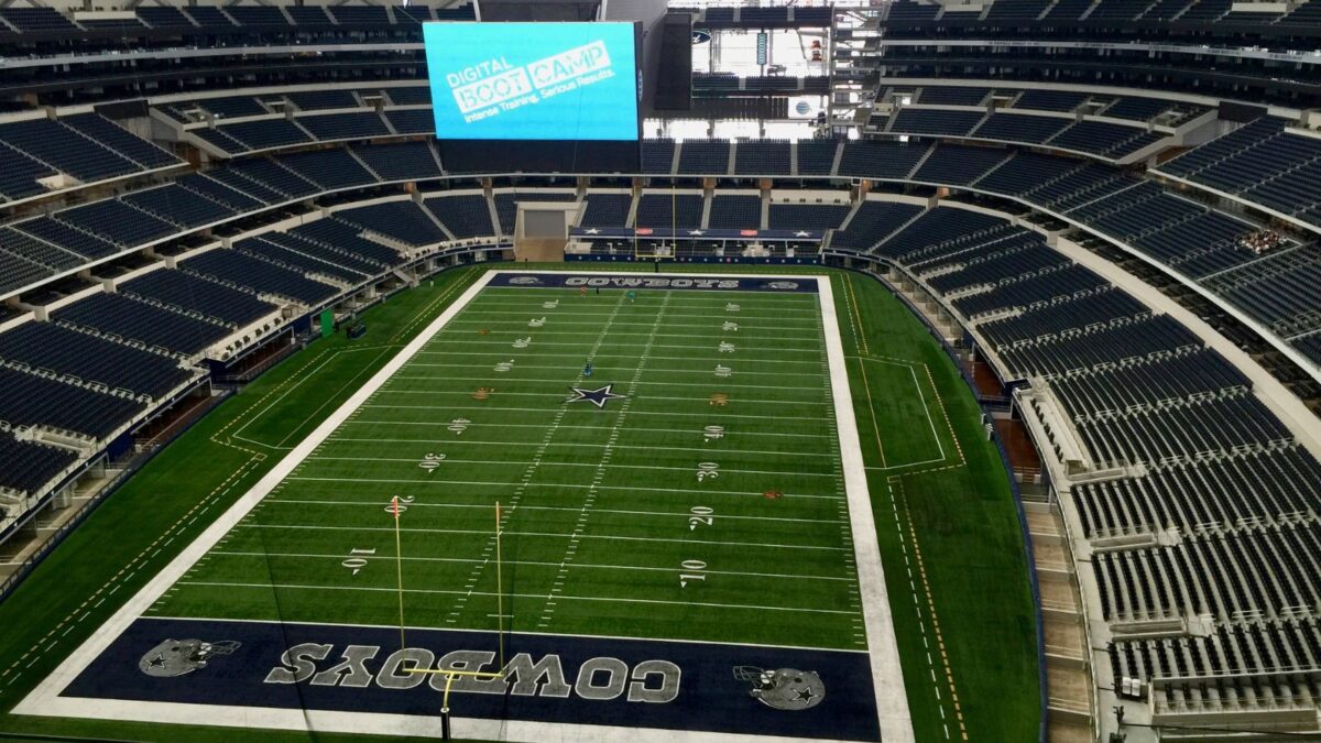 10 Iconic Football Stadiums Every Fan Should Experience 4 The vast interior of AT&T Stadium, home of the Dallas Cowboys, with the playing field covered by a large digital screen displaying the message "Digital Bootcamp." The stands are empty, and the stadium's immense size is highlighted by the rows upon rows of seating and the gigantic overhead screen.