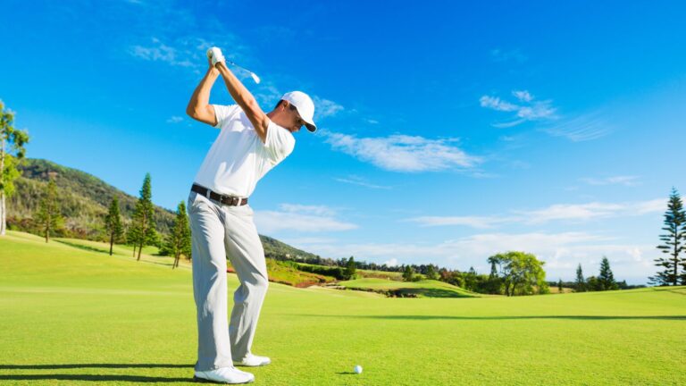 Golfer in mid-swing on a sunny, lush golf course with a clear blue sky in the background.