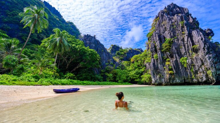 A person wading in the clear shallow waters of a secluded beach, with a kayak on the sandy shore and towering limestone cliffs surrounded by lush greenery under a blue sky with scattered clouds.