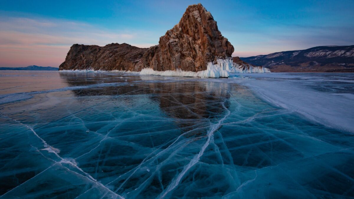 20 Breathtaking Lakes That Are True Natural Wonders! 12 A solitary rock formation rises from the frozen surface of Lake Baikal, showcasing the clear blue ice and distant mountains at twilight.