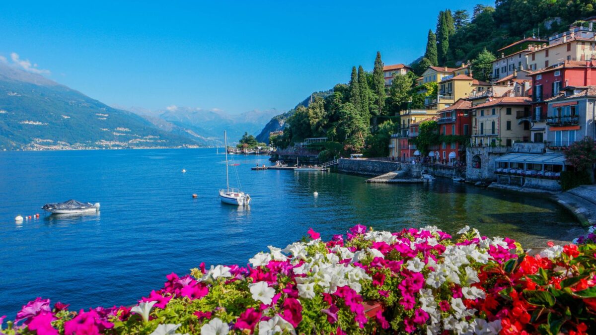 20 Breathtaking Lakes That Are True Natural Wonders! 13 A picturesque view of colorful houses lining the shore of Lake Como, with blooming flowers in the foreground and mountains in the distance.