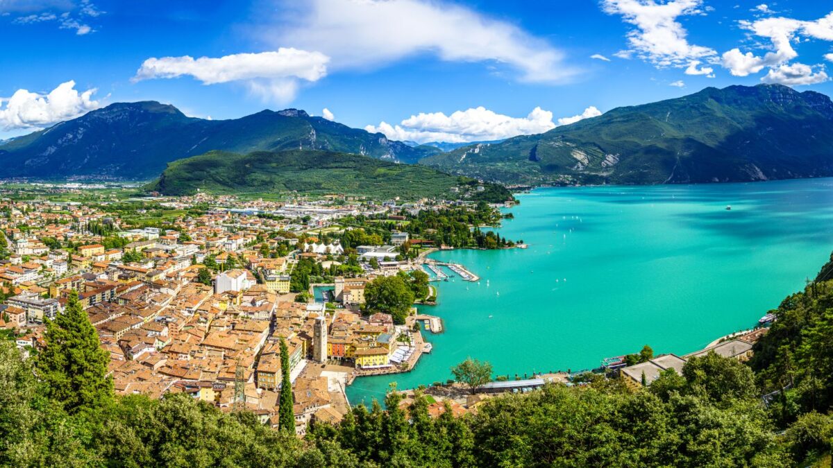 20 Breathtaking Lakes That Are True Natural Wonders! 18 A panoramic view of Lake Garda, showcasing the bustling lakeside town and the vast, tranquil waters against a backdrop of mountains and blue skies.