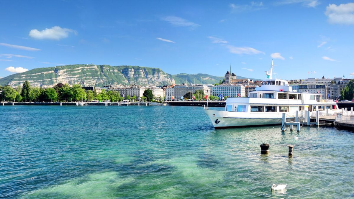 20 Breathtaking Lakes That Are True Natural Wonders! 20 The clear waters of Lake Geneva with a view of the cityscape and a large hill in the background, a boat docked in the foreground, and a swan swimming by.