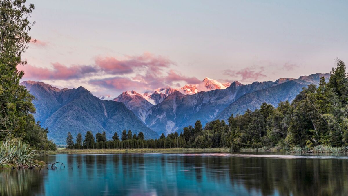 20 Breathtaking Lakes That Are True Natural Wonders! 22 Dusk at Lake Matheson, with calm waters reflecting the pink-tinged snow-capped mountains and lush greenery.