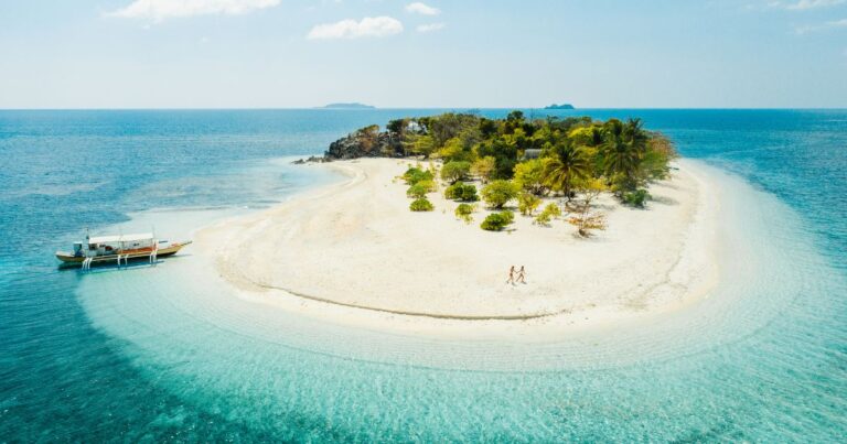 A couple walking on a secluded white sand beach with a boat moored in clear blue water.