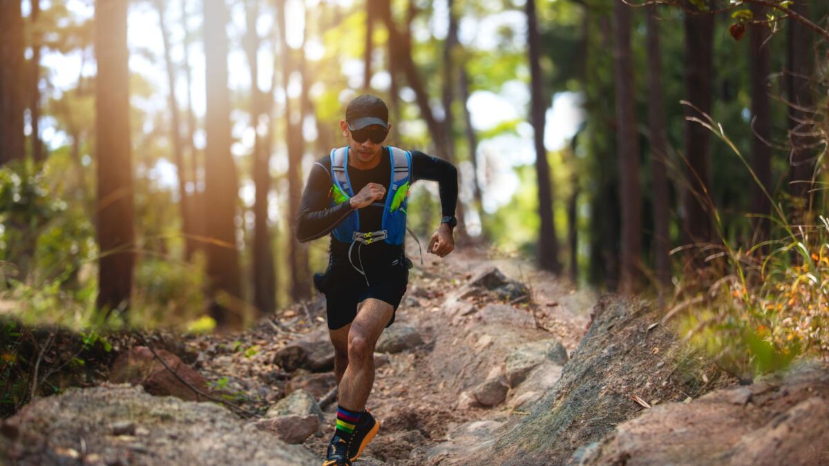 A trail runner, equipped with a hydration pack, is racing along a rugged, rocky trail through a sun-dappled forest. The athlete is wearing a black visor, sunglasses, and a focused expression as they navigate the challenging terrain.