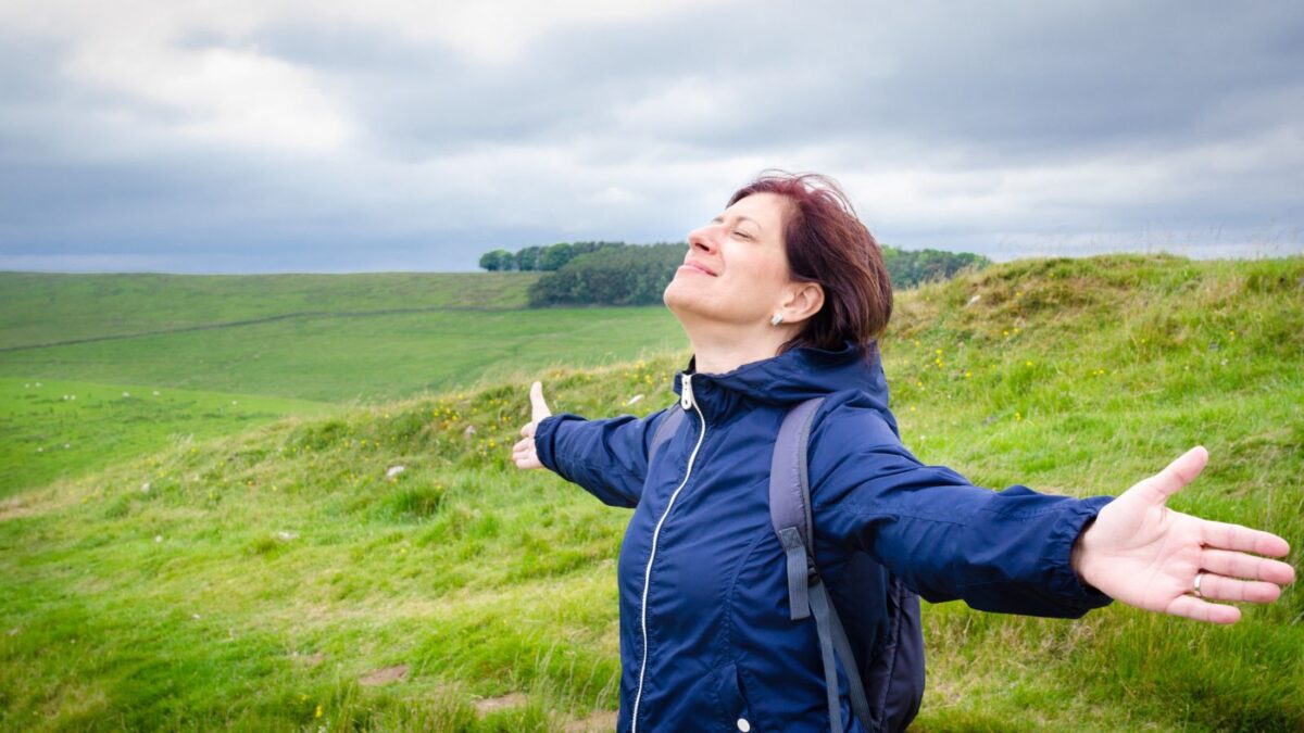 A woman in a navy blue jacket with arms outstretched is inhaling deeply, enjoying the fresh air and freedom atop a green, windy hillside with sparse trees and a grey sky above, embodying a sense of peace and contentment.